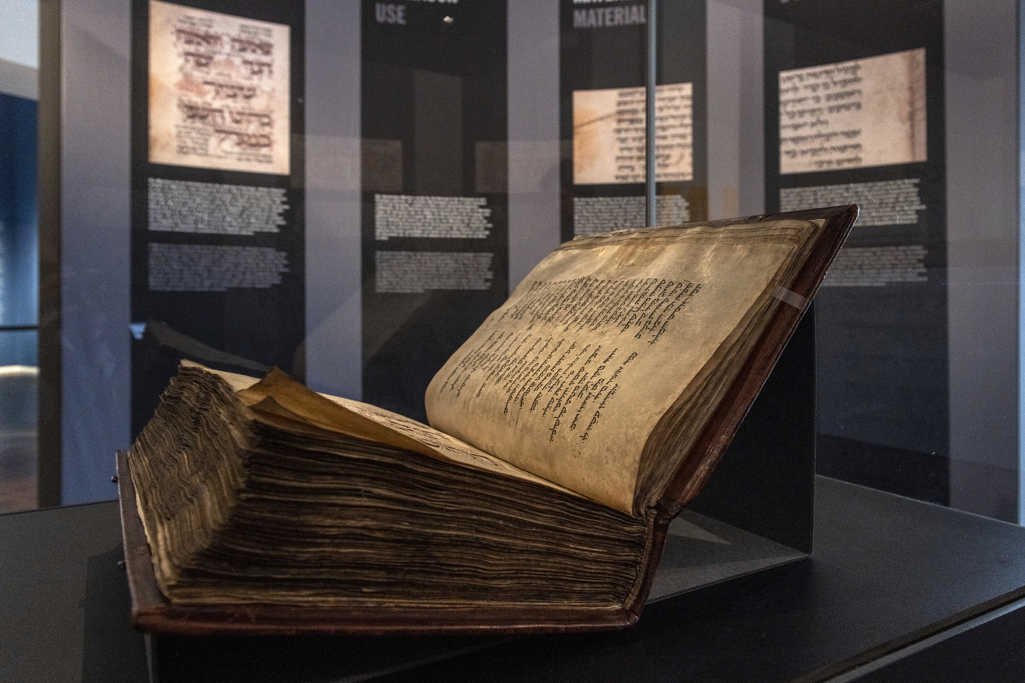 Der Amsterdam Machsor, ein dickes, altes Buch aus dem Mittelalter, liegt aufgeklappt in einer Vitrine, die sich in einem Ausstellungsraum im Wallraf-Richartz-Museum befindet. Die aufgeklappte Buchseite zeigt hebräische Schrift. Im Hintergrund eine dunkle Wand mit Ausstellungstexten.