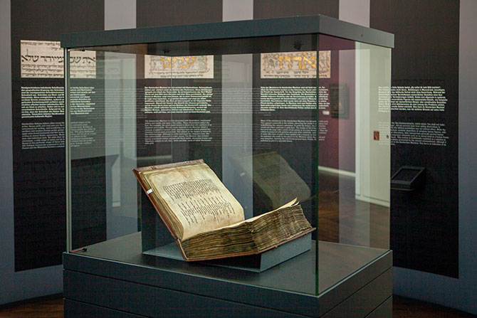 Der Amsterdam Machsor in einer Vitrine im Wallraf-Richartz-Museum. Das alte Buch ist aufgeklappt. Hinter der Vitrine sind dunkle Ausstellungswände, die Erklärtexte präsenieren.