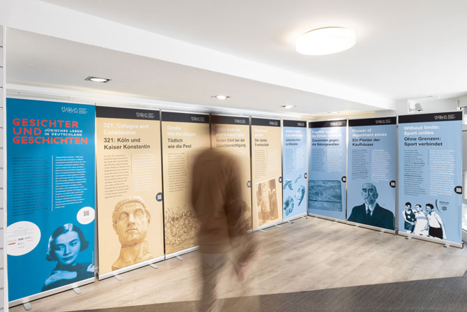 Frontansicht der Roll-Ups der Posterausstellung. Ein Mann bewegt sich vor der Posterausstellung, seine Bewegung ist verschwommen. Die Posterausstellung im Hintergrund ist scharf gestellt.