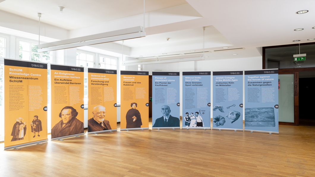 Die Posterausstellung ist halbrund in einem Innenraum des Zentrums für verfolgte Künste aufgestellt. Der Boden ist aus Holz. Links im Bild sind vier gelbe Roll-Ups aufgestellt, daran angrenzend sind rechts vier blaue Roll-Ups aufgestellt.