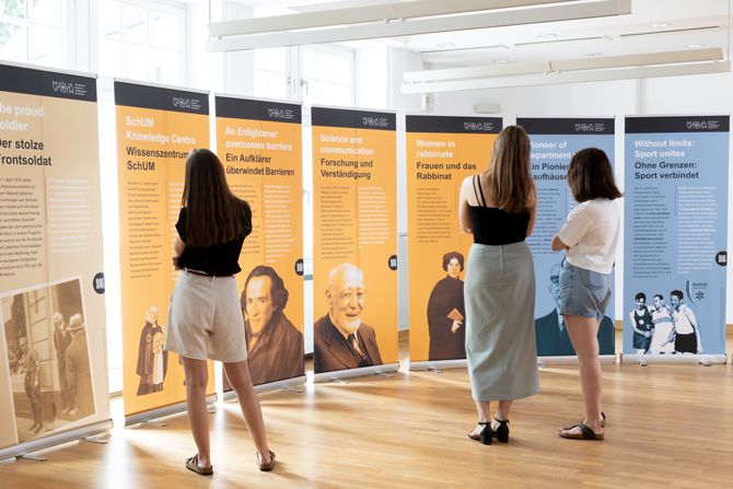 Die Posterausstellung ist in einem Raum mit Holzboden des Zentrum für verfolgte Künste aufgestellt. Drei Frauen stehen vor den Roll-ups und sind von hinten fotografiert.