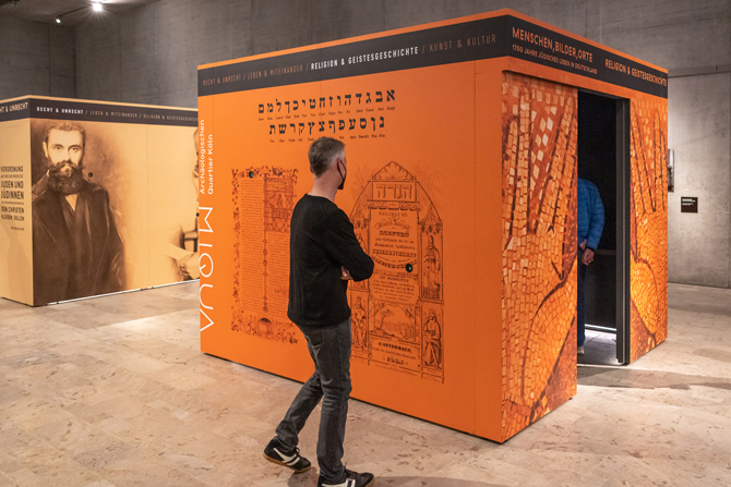 Ein Mann steht vor dem orangenen Ausstellugnskubus zum Thema "Religion und Geistesgeschichte". Links im Hintergrund ist der gelbe Ausstellungskubus zu erkennen.