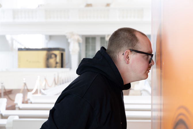 Ein Mann mit Brille schaut von außen durch ein Guckloch hinein in einen orangenen Ausstellungskubus, der in der Ludwigkirche Saarbrücken steht.