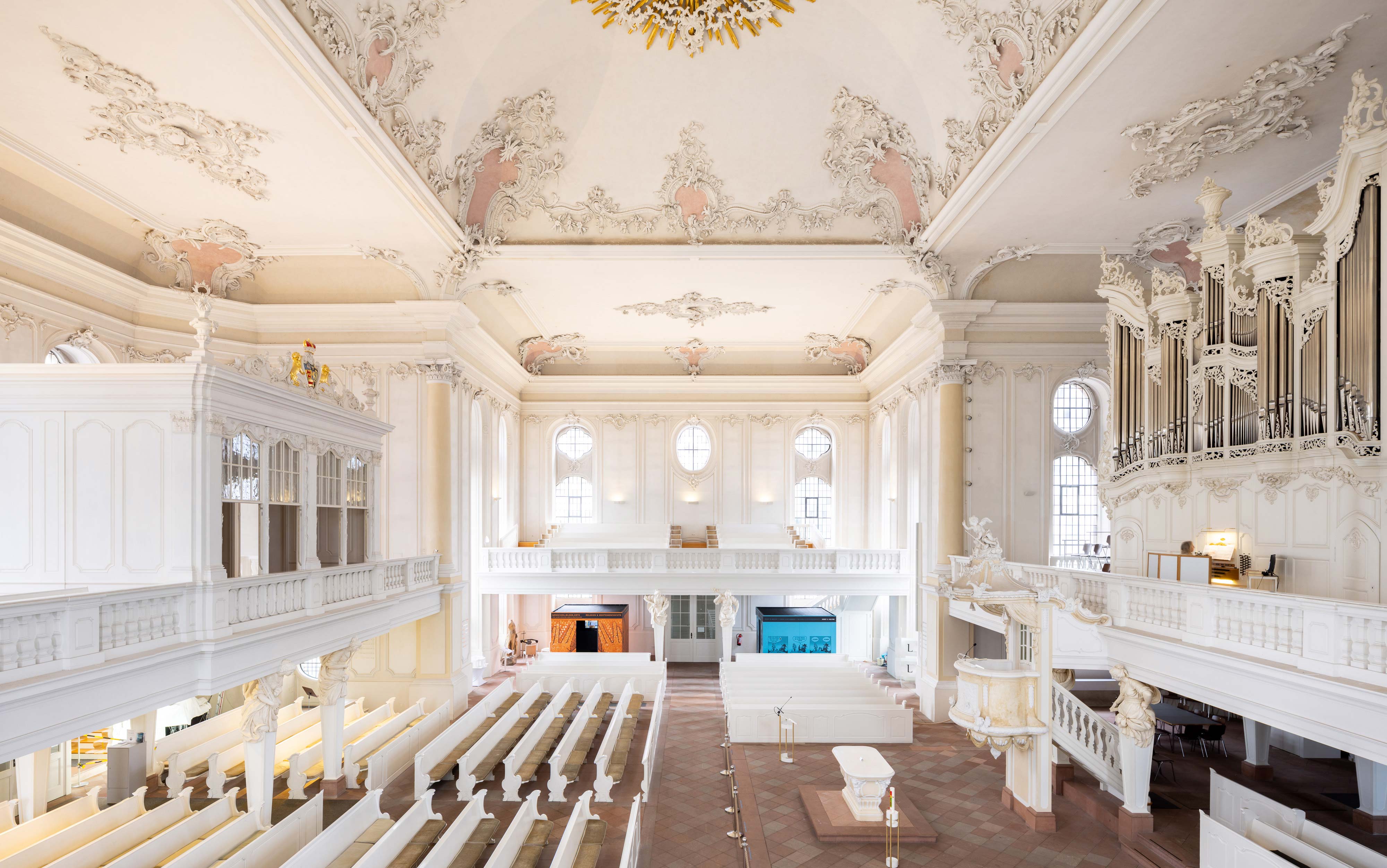 Blick von einer Empore in der Ludwigskirche in Saarbrücken hinein in den großen Raum der hellen Kirche. Im Erdgeschoss der Kirche sind zwei Ausstellungskuben platziert.