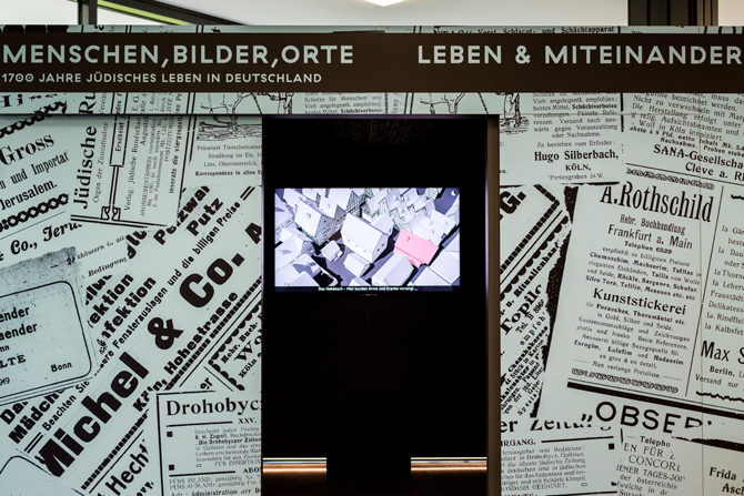 Frontansicht des Ausstellungskubus zum Thema "Leben & Miteinander". In dem Kubus ist ein Screen zu sehen, der Häuser zeigt. 