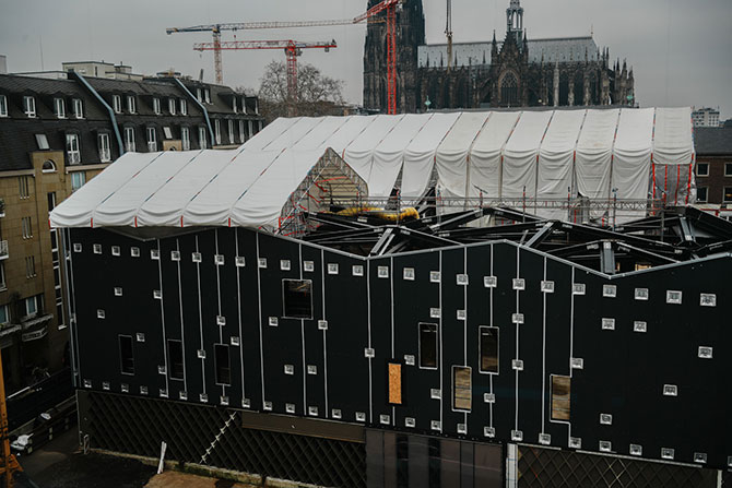 Eine Baustelle ist komplett mit einer schwarzen Stahlstruktur bebaut.    Das Dach besteht aus einem Pyramiden-artigen Stahlkonstrukt, dass zum Teil mit einer weißen Plastikfolie bedeckt ist.  Die Fassade des Gebäudes ist dunkelgrau.