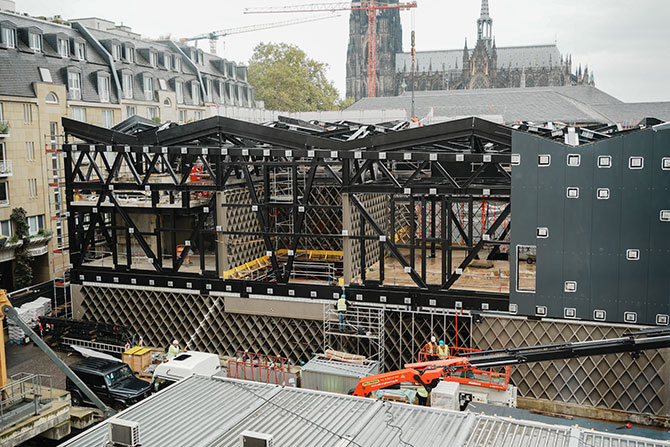 Eine Baustelle ist komplett mit einer schwarzen Stahlstruktur bebaut.  Im unteren Stockwerk sind Rautentragwerke eingebaut.  Das Dach besteht aus einem Pyramiden-artigen Stahlkonstrukt.  Zum Teil ist eine graue Fassade zu sehen.