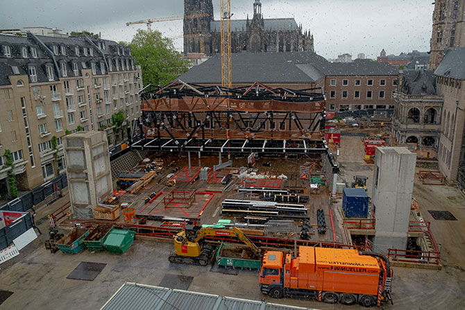 Eine Baustelle die von historischen Häusern umringt ist.  In der Mitte ist ein Teil der Baustelle mit einer schwarzen Stahlstruktur bebaut.  Links und rechts vorne sind zwei graue Betontürme zu sehen.  Vorne im Bild ist ein orange-farbiger LKW zu sehen.