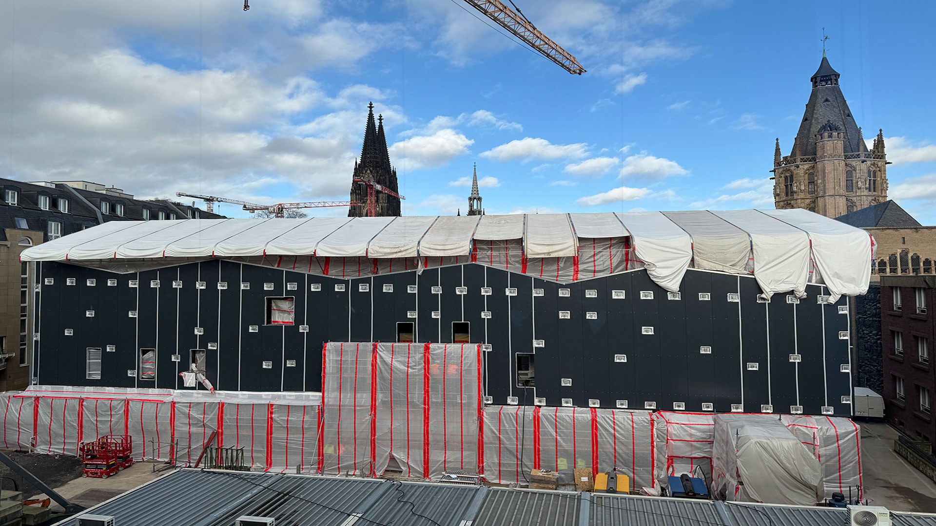 Eine dunkles, zweistöckiges Gebäude ist zu sehen, dass noch im Bau ist.  Ein weißer Bauzaun steht um die komplette Baustelle.  Das Dach ist mit einer weißen Plastikfolie bedeckt.