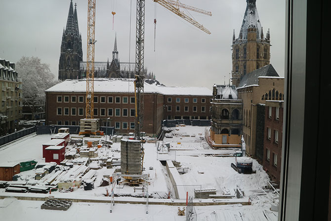 Eine Baustelle ist zu sehen, in deren Mitte zwei große Kräne stehen.  Umgeben ist sie von historischen Häusern.  Im Hintergrund sieht man eine große Kirche.  Alles ist mit Schnee bedeckt.