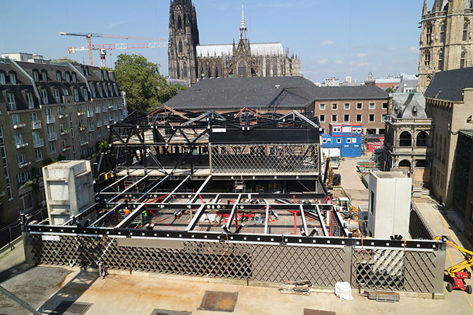 Eine Baustelle die von historischen Häusern umringt ist.  In der Mitte ist ein Teil der Baustelle mit einer schwarzen Stahlstruktur überbaut.  Eine Stahlwand aus Rauten ist im oberen Stockwerk als Wand eingesetzt. Links und rechts vorne sind zwei graue Betontürme zu sehen.  