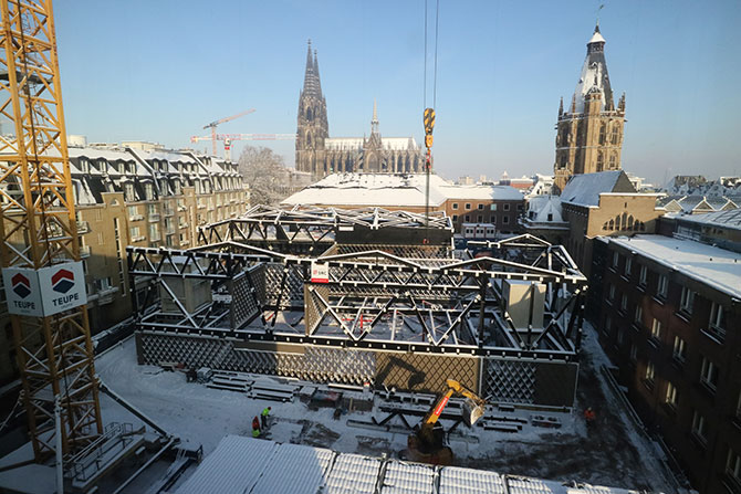 Eine Baustelle die von historischen Häusern umringt ist. Die Baustelle ist komplett mit einer schwarzen Stahlstruktur bebaut. Im unteren Stockwerk sind Rautentragwerke eingebaut. Links und rechts vorne sind zwei graue Betontürme zu sehen. Die ganze Baustelle ist mit Schnee bedeckt.