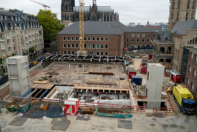 Eine Baustelle, die mit einer Holzstruktur überlegt ist.  Links und rechts der Baustelle sind zwei kleine Türme aus Beton zu sehen. Im hinteren Teil der Baustelle sieht man einen Kran sowie eine schwarze Stahlstruktur.