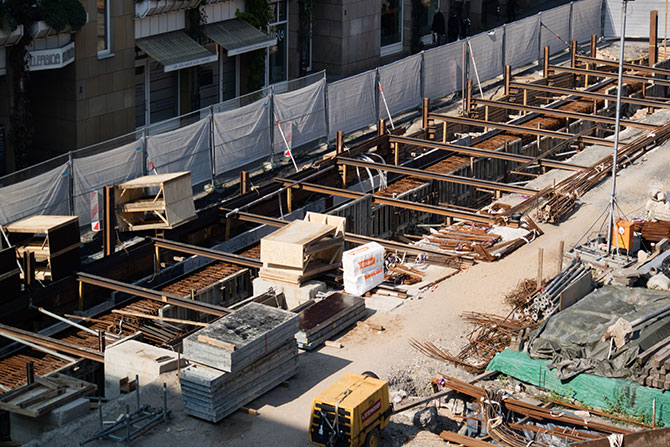 Eine Baustelle, die durch Sichtfolien abgeschirmt ist.  Der Blickwinkel ist von oben, von einem anderen Gebäude aus. Man kann eine ausgehobene Tranche sehen, die durch Stahl gestützt wird.