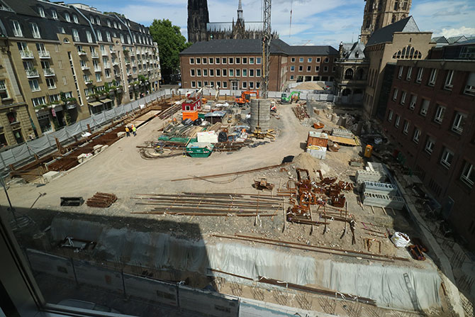 Eine Baustelle ist zu sehen.  Viel Material wie Stahlstangen liegen auf der Erde.  Um die Baustelle rum sind historische Gebäude zu sehen.