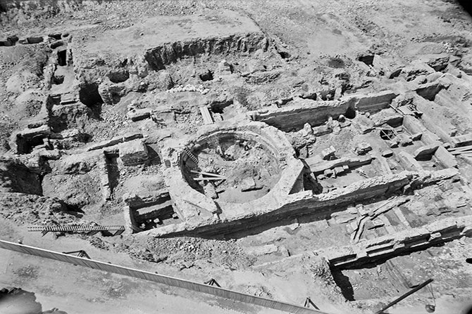 Schwarz-Weiß-Foto einer archäologischen Ausgrabung – Die Aufnahme zeigt die freigelegten Überreste einer historischen Gebäudestruktur. In der Mitte ist eine kreisförmige Mauer zu erkennen, umgeben von rechteckigen Fundamenten und Mauern aus Stein. Der Boden ist uneben und von Erdschichten durchzogen. Verschiedene Gräben, freigelegte Mauerreste und Werkzeuge der Archäologen deuten auf eine laufende Untersuchung hin.