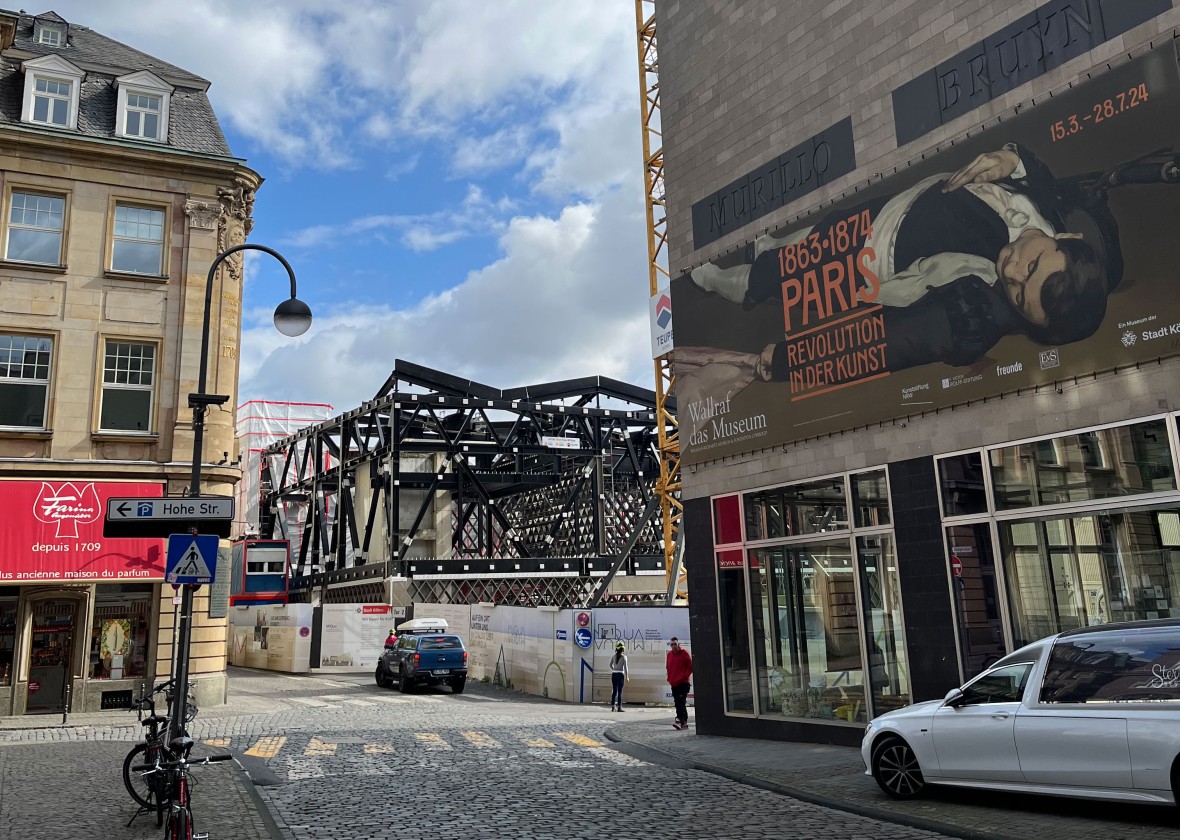 Ein Blick auf die MiQua-Baustelle. Die Stahlträger des Museumsneubaus stehen bereits. Rechts im Bild befindet sich das Eckgebäude des Wallraf-Richartz-Museums. Links im Bild ist das Altbaugebäude, in dem sich im Erdgeschoss das Farina-Duftmuseum befindet. Der Himmel ist blau und bewölkt..