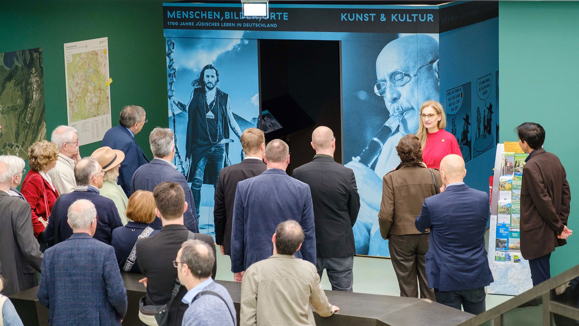 Besucher*innen werden von einer Kuratorin durch die Wanderausstellung "Menschen, Bilder Orte - 1700 Jahre jüdisches Leben in Deutschland" geführt. Die Kuratorin steht vor einem blauen, begehbaren Ausstellungskubus. 