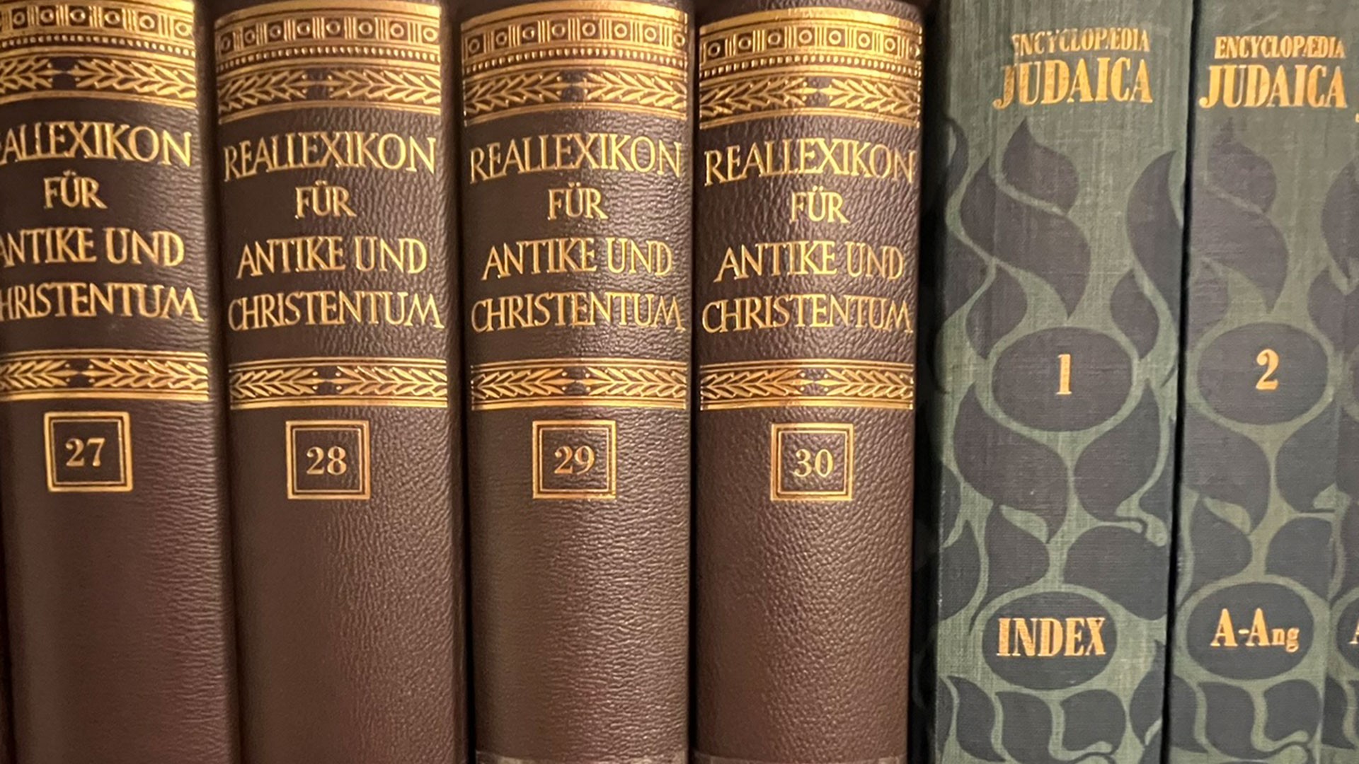 Nahaufnahme einer Sammlung von alten Büchern, dessen Rücken man sehen kann. Links im Bild sind vier braune Bücher mit goldener Verzierung. Rechts im Bild zwei grau-blaue Bücher, ebenfalls mit goldener Verzierung.