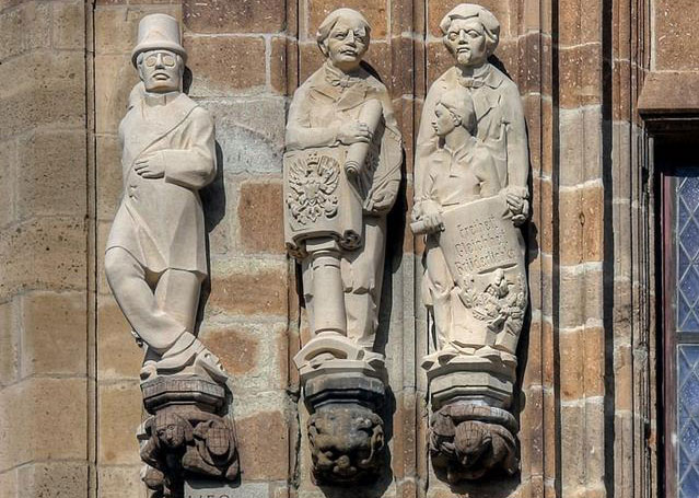 Drei Steinfiguren am Rathausturm in Köln. Die Figur links trägt eine runde Brille und einen Zylinderhut. Die Figur in der Mitte trägt eine Rolle. Die Figur rechts im Bild hat eine weitere, kleinere Figur vor sich und trägt eine Rolle, auf der "Freiheit, Gleichheit, Brüderlichkeit" steht. 