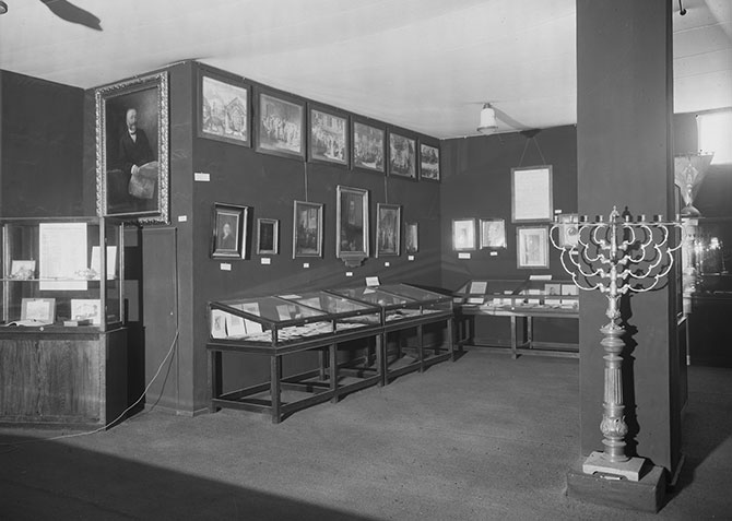 Einblick in die Jahrtausend-Ausstellung der Rheinlande mit der Abteilung "Juden und Judentum im Rheinland". Im Raum stehen verschiedene Vitrinen, in denen Judaica ausgestellt werden. An den Wänden hängen verschiedene Bilder. Im Vordergrund steht eine Menora an einer Säule. 