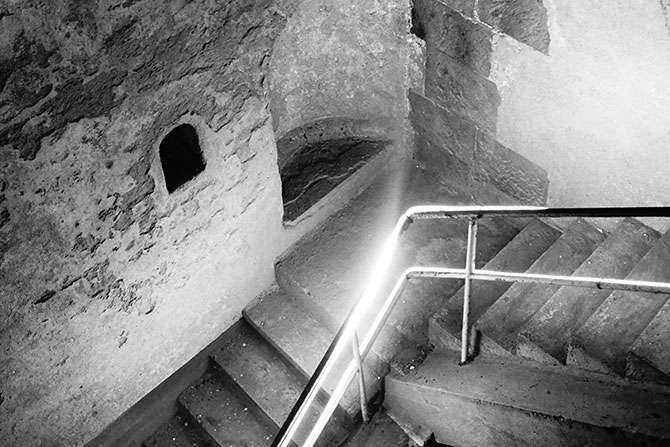 Eine Schwarzweiß-Fotografie des Eingangs in der Kölner Mikwe im unterirdischen Rundgang von MiQua. Die Treppen runter zur Mikwe sind in einer Vogelperspektive fotografiert. Die Mauern im Treppengang zur Mikwe zeigen eine Nische für ein Fenster sowie eine weitere, größere Nische.