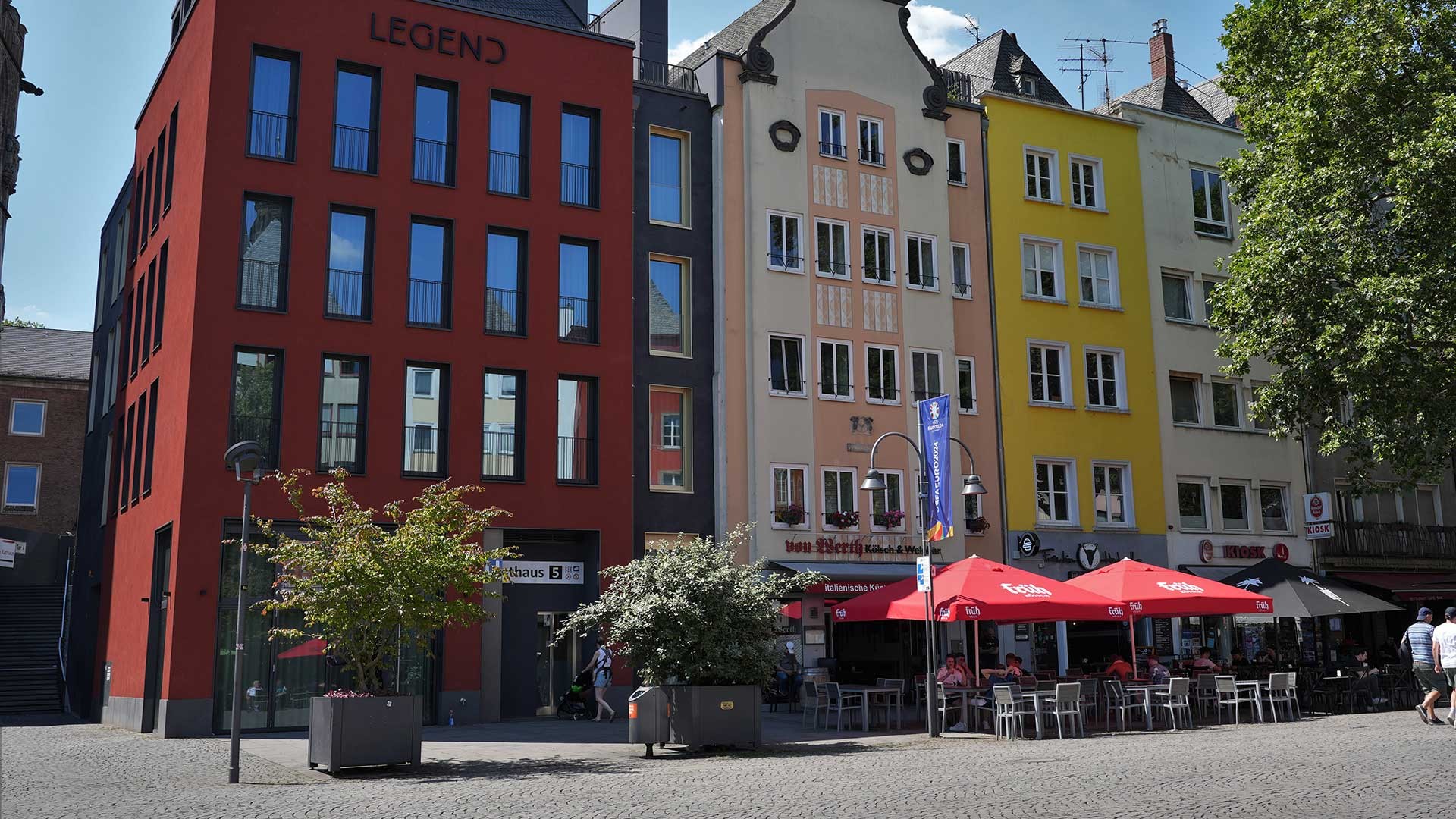 Vier Häuser am Alter Markt in Köln. Links im Bild das Rote Haus, in dem sich das MiQua:forum unten im Erdgeschoss befindet. Rechts daneben befinden sich drei weitere, schmale Häuser. Vor den Häusern stehen Bäume und zwei rote Pavillons.