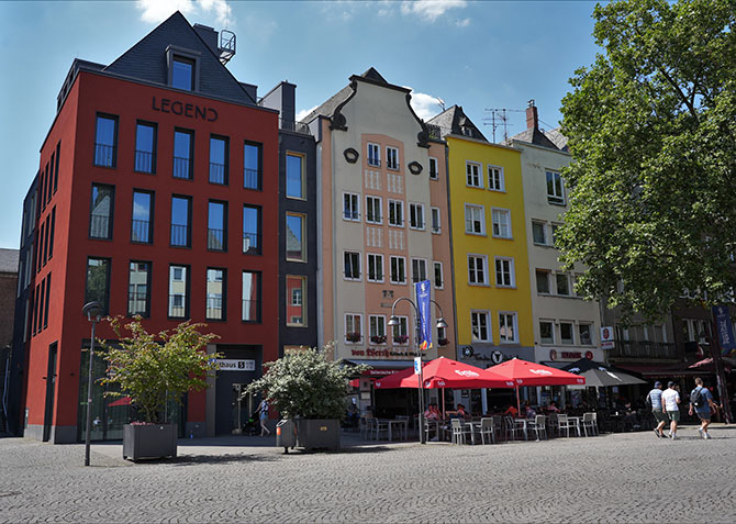 Ein rotes Haus in der Altstadt von Köln. Hier befindet sich das MiQua.forum im Erdgeschoss. An das Rote Haus grenzt rechts ein rosa, ein gelbes und ein weißes Haus. 