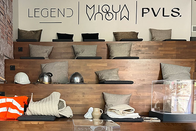 Eine Ansicht des MiQua:forum mit Sitzflächen für Gäste. Auf den Sitzflächen liegen Kissen. Auf der ersten Stufe ist eine Vitrine angebracht. Oben an der Rückwand hängen die Logos von Legend, MiQua und Pvls.