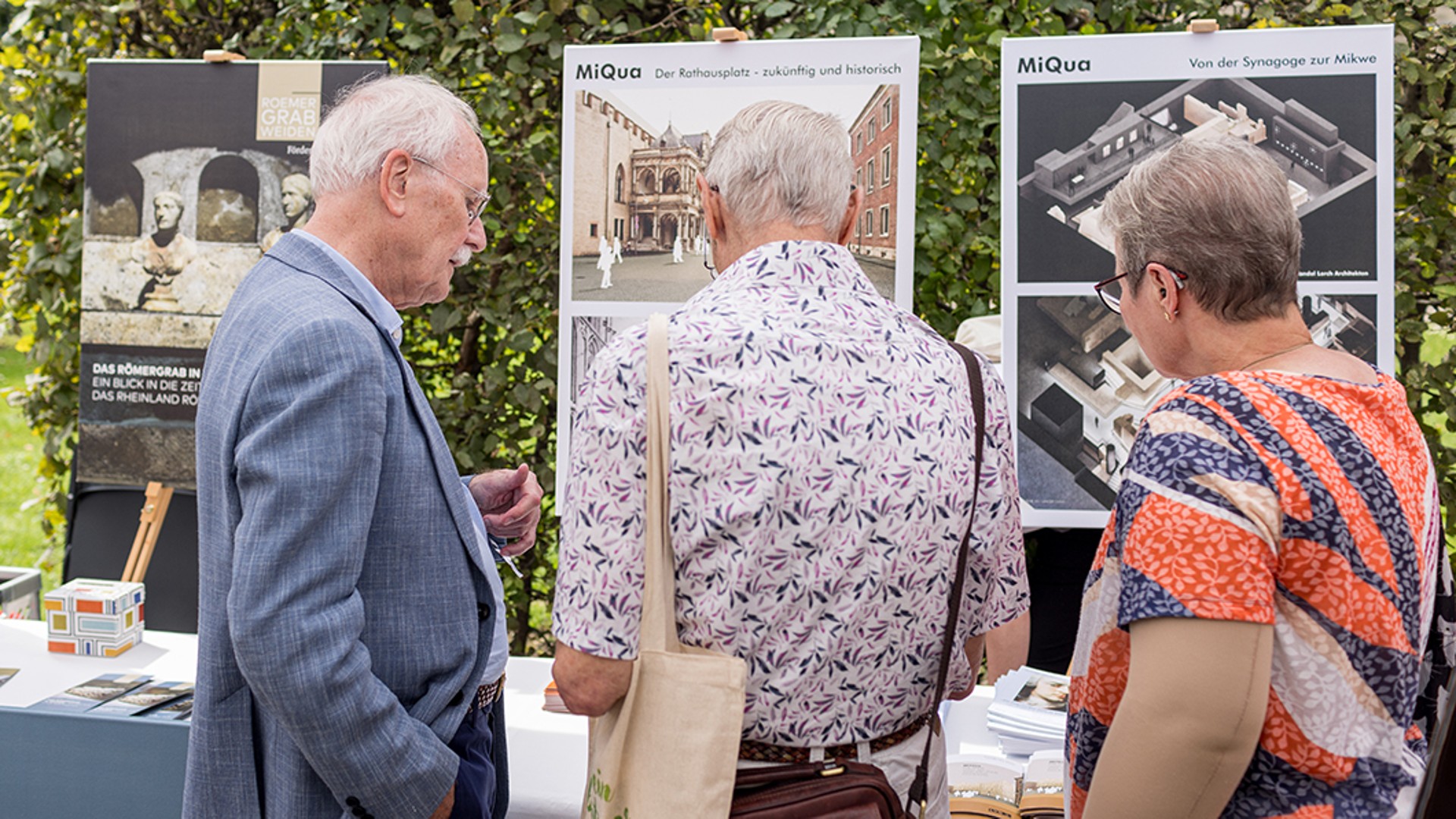 Zwei Männer und eine Frau stehen mit dem Rücken zur Kamera.  Sie betrachten ein Roll-up der MiQua-Freunde.