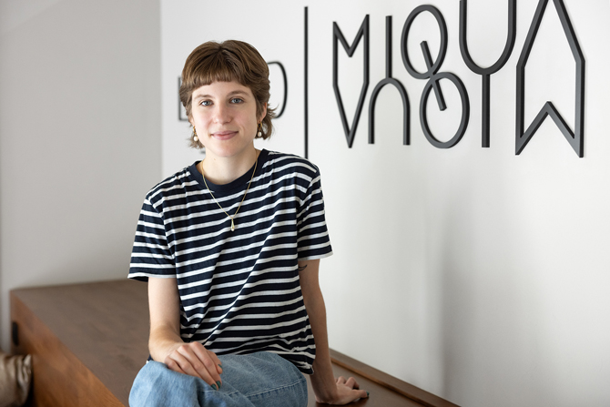 Eine jüngere Mitarbeiterin mit kurzen brauen Haaren trägt ein gestreiftes T-Shirt und blaue Jeans und sitzt auf einer Holztreppe. Im Hintergrund das Logo von MiQua an einer weißen Wand. 