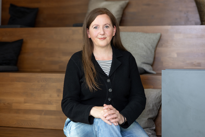 Portrait einer Mitarbeiterin von MiQua, die auf einer Holztreppe sitzt und in die Kamera schaut. Sie hat braune lange Haare, trägt eine schwarze Strickjacke, ein gestreiftes Oberteil und eine blaue Jeans. 