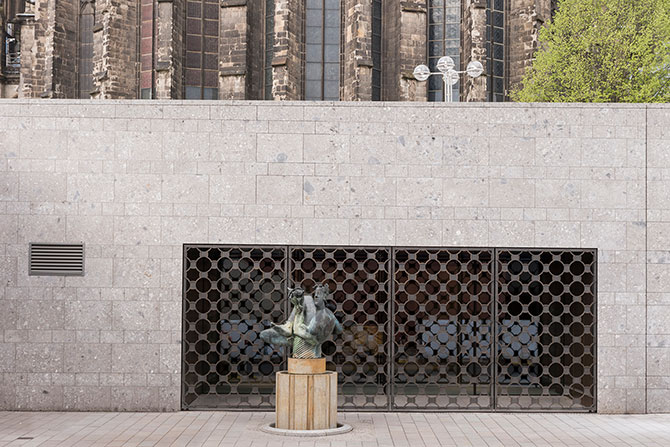 Eine Wand aus Stein ist zu sehen. In der Mitte ist eine Öffnung, die durch eine rautenförmige, durchsehbare Abdeckung aus Metall verschlossen ist. Im Vordergrund ist ein Podest mit einer Statue zu sehen.