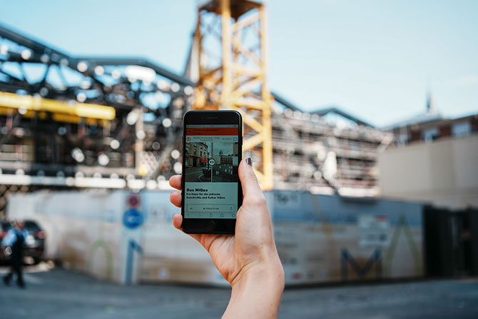 Im Hintergrund ist eine Baustelle zu sehen, die von einem Bauzaun umringt ist. Im Vordergrund ist eine Hand zu sehen, die ein Smartphone in die Luft hält. Auf dem Handy ist eine App zu sehen.