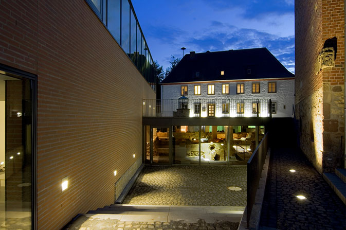 Ein zweistöckiges, älteres, weißes Haus ist im Hintergrund zu erkennen. Die Fenster sind hell beleuchtet, der Himmel ist dunkel. Vor dem Haus ist ein moderner Glasanbau zu sehen, der ebenfalls mit einem warmen Licht beleuchtet ist. Links im Bild ist eine lange, gelbe Wand zu sehen. Rechts im Bild ist die Fassade eines älteren Haus zu sehen. Bodenleuchter strahlen von unten warmes Licht entlang der Wand.