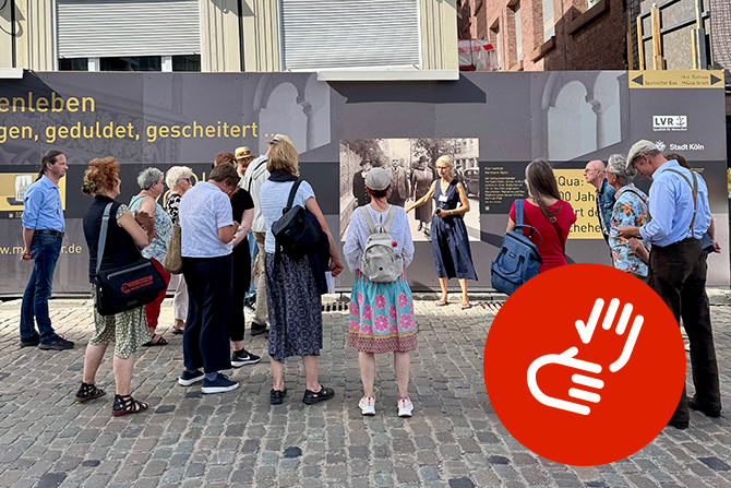 Eine Gruppe von Menschen steht auf einem Kopfsteinpflasterplatz vor einer Bauzauninstallation mit Infotafeln und Bildern. Eine Frau, die offenbar als Führerin auftritt, spricht zu den Zuhörenden und deutet dabei mit einer Geste. Die Teilnehmenden hören aufmerksam zu, einige tragen Rucksäcke oder Taschen. Auf dem Bauzaun sind große Texte und historische Fotos zu sehen. Die Szene vermittelt den Eindruck einer Stadtführung oder einer historischen Exkursion.