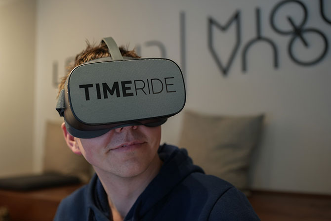 Ein junger Mann trägt eine Virtual Reality Brille auf der "TimeRide" steht und schaut damit in die Kamera.