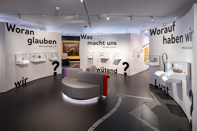 Ein Blick in eine Ausstellung. Der Raum ist rund. An den Wänden stehen Worte wie "Woran glauben wir?" und "Was macht uns wütend?" An den Wänden hängen kleine Vitrine die Ausstellungstücke beinhalten. Mitten im Raum steht ein rundes, graues Sofa.
