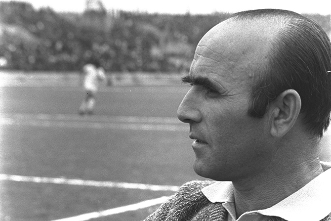 Ein schwarz-weiß Foto. Der Kopf eines Mannes mit leichter Glatze ist von der Seite zu sehen. Im Hintergrund ist ein Fußballfeld zu erkennen.