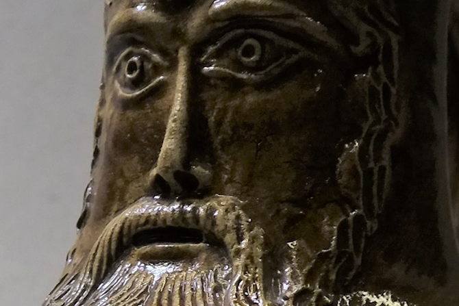 Ein dunkelbrauner Krug aus Ton, auf dem das Gesicht eines bärtigen Manns zu sehen ist.