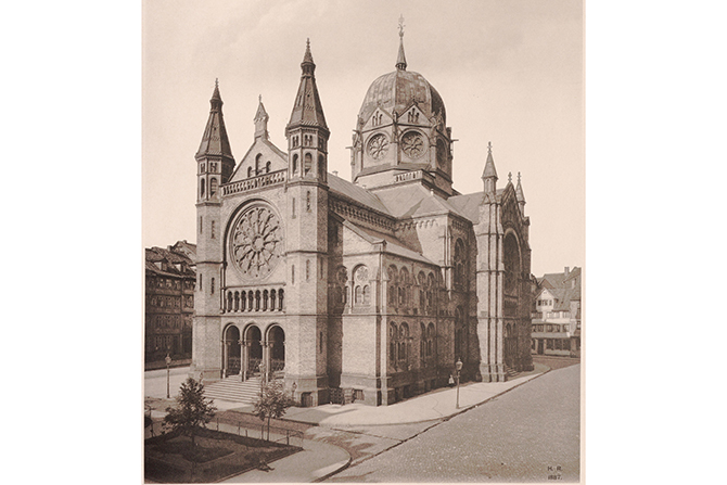 Das Foto zeigt ein großes, monumentales Gebäude im Stil historistischer Architektur. Es besitzt einen hohen, reich verzierten Kuppelbau in der Mitte, flankiert von zwei schlanken Türmen mit spitzen Helmen. Die Fassade ist geprägt von einem großen Rundfenster mit ornamentalen Glaseinsätzen, das oberhalb eines Säulenportals mit drei Eingängen liegt. Das Bauwerk ist aus Stein errichtet und weist zahlreiche dekorative Elemente wie Arkaden, Gesimse und filigrane Verzierungen auf. Rechts und links schließen sich weitere Gebäudeteile mit kleineren Türmen und Fenstern an. Im Vordergrund ist eine Straße mit Bäumen und Gehwegen erkennbar, während im Hintergrund weitere Wohnhäuser stehen.