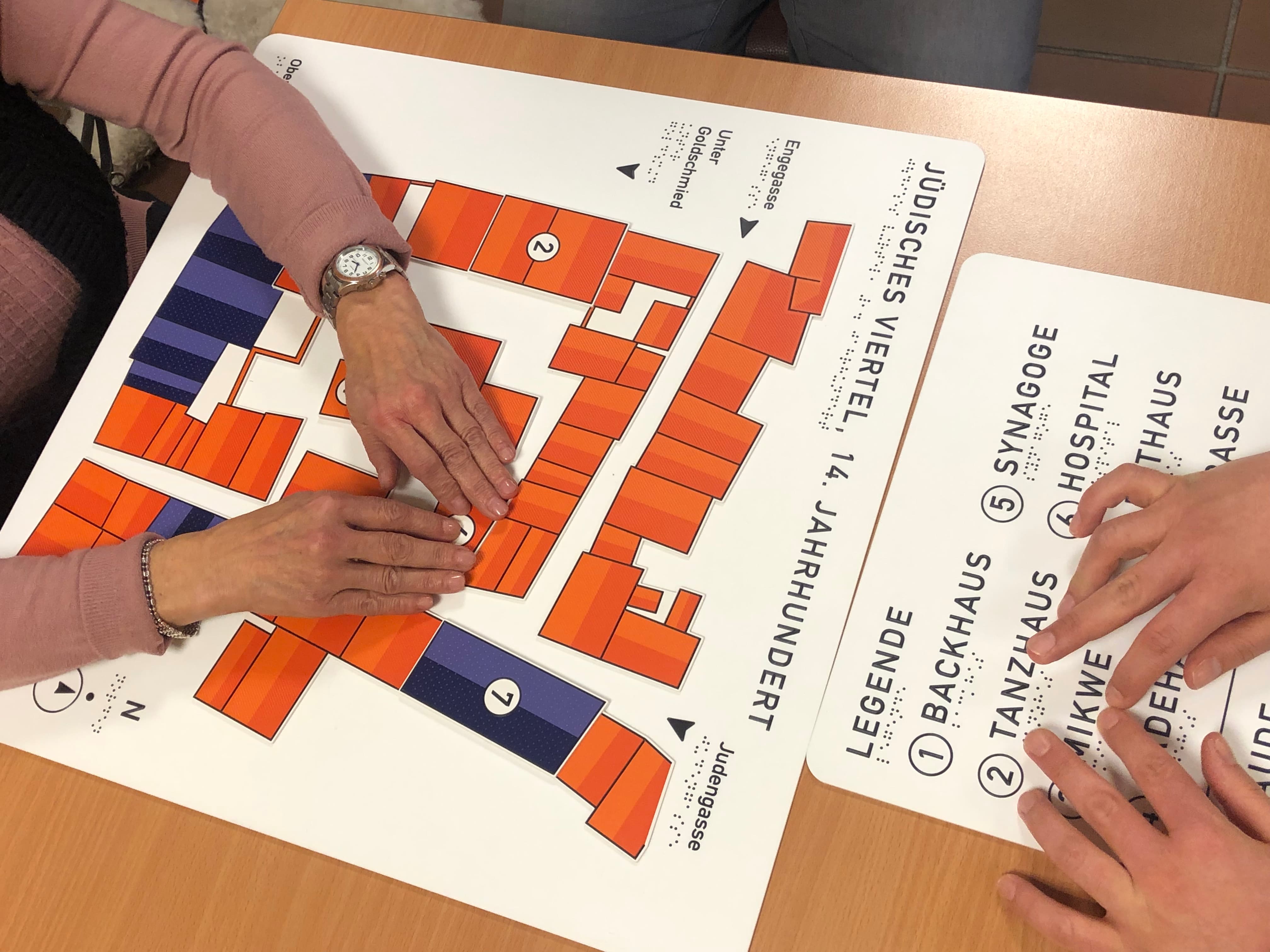 Zwei paar Hände liegen auf einem Tastplan mit orangenen und blauen Rechtecken und Blindeschrift.
