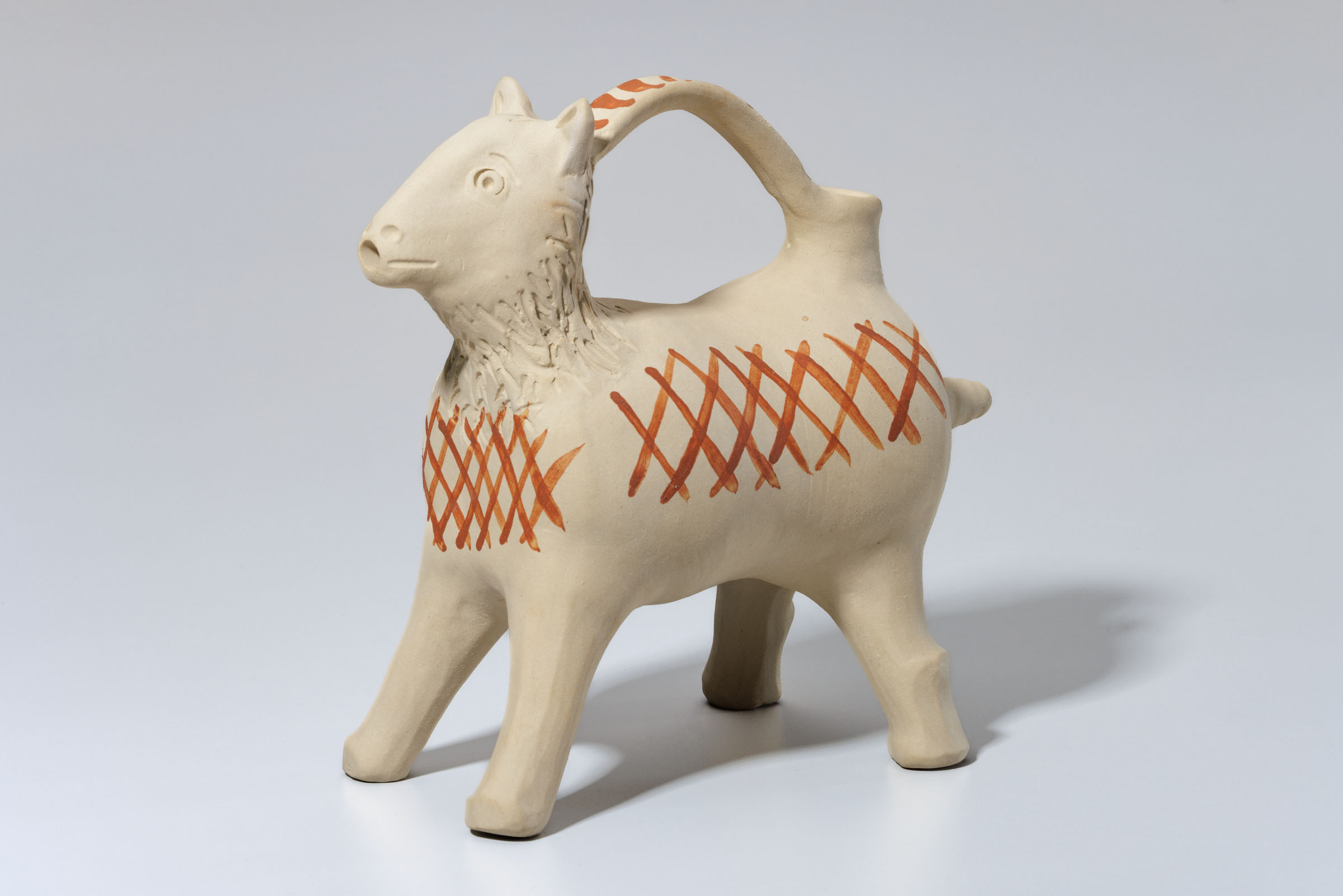 Ein weißes Tongefäß mit roten Streifen die sich kreuzen. Das Gefäß hat die Form eines Hundes oder einer Ziege und einen Henkel oben, an dem man es festhalten kann.
