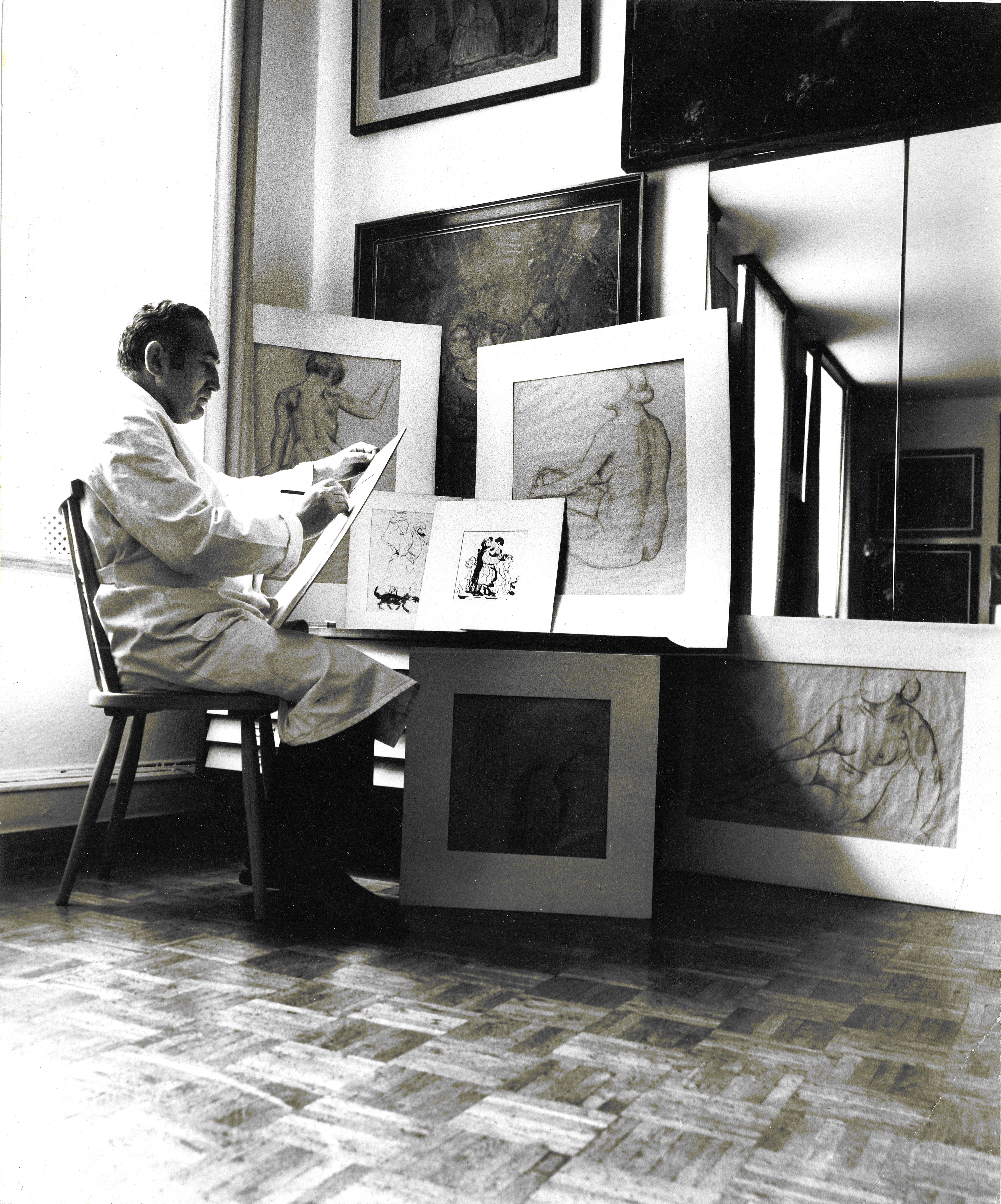 Ein schwarz-weiß Foto eines Manns in einem weißen Kittel. Er sitzt auf einem Stuhl und zeichnet mit einem Stift auf ein großes Papier. Vor ihm stehen und hängen mehrere Aktzeichnungen von Frauen.