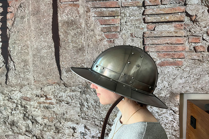 Eine Frau trägt einen runden Helm aus Stahl vor einer Mauer aus Stein.