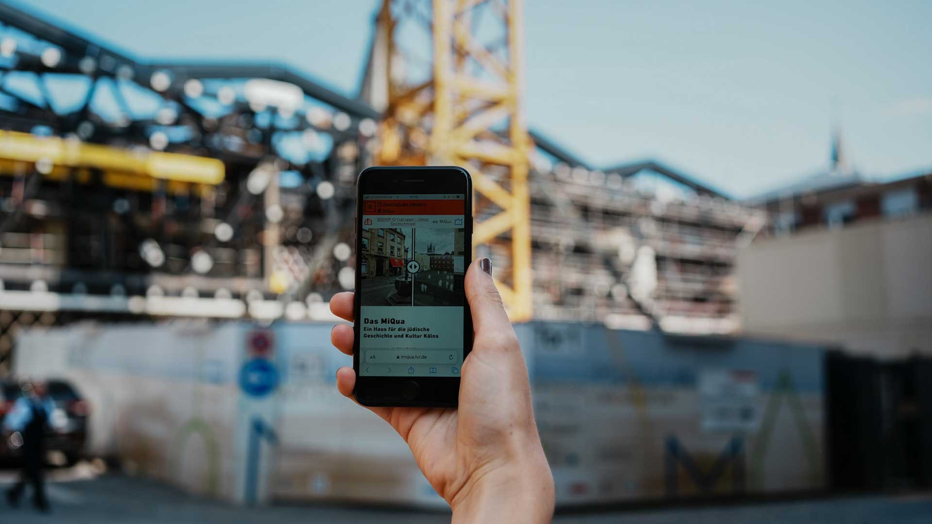 Eine Hand hält ein Smartphone hoch.  Im Hintergrund ist die Baustelle von MiQua zu sehen.  Auf dem Smartphone ist die App Zwischen den Häusern zu sehen.