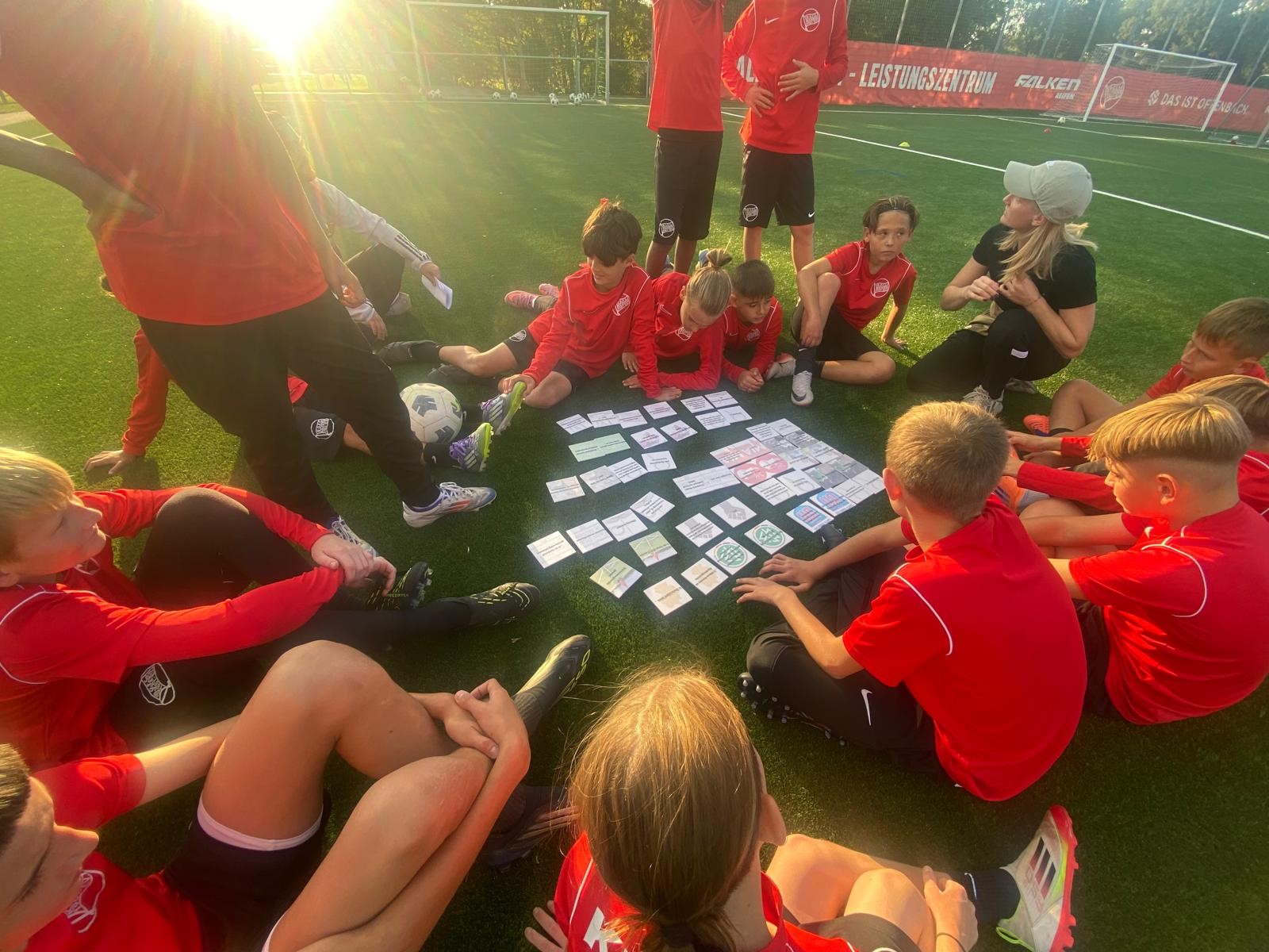 Eine Gruppe von Kindern in roten T-Shirts sitzen im Kreis auf einem Fußballfeld. In der Mitte liegen weiße Karten auf dem Boden.