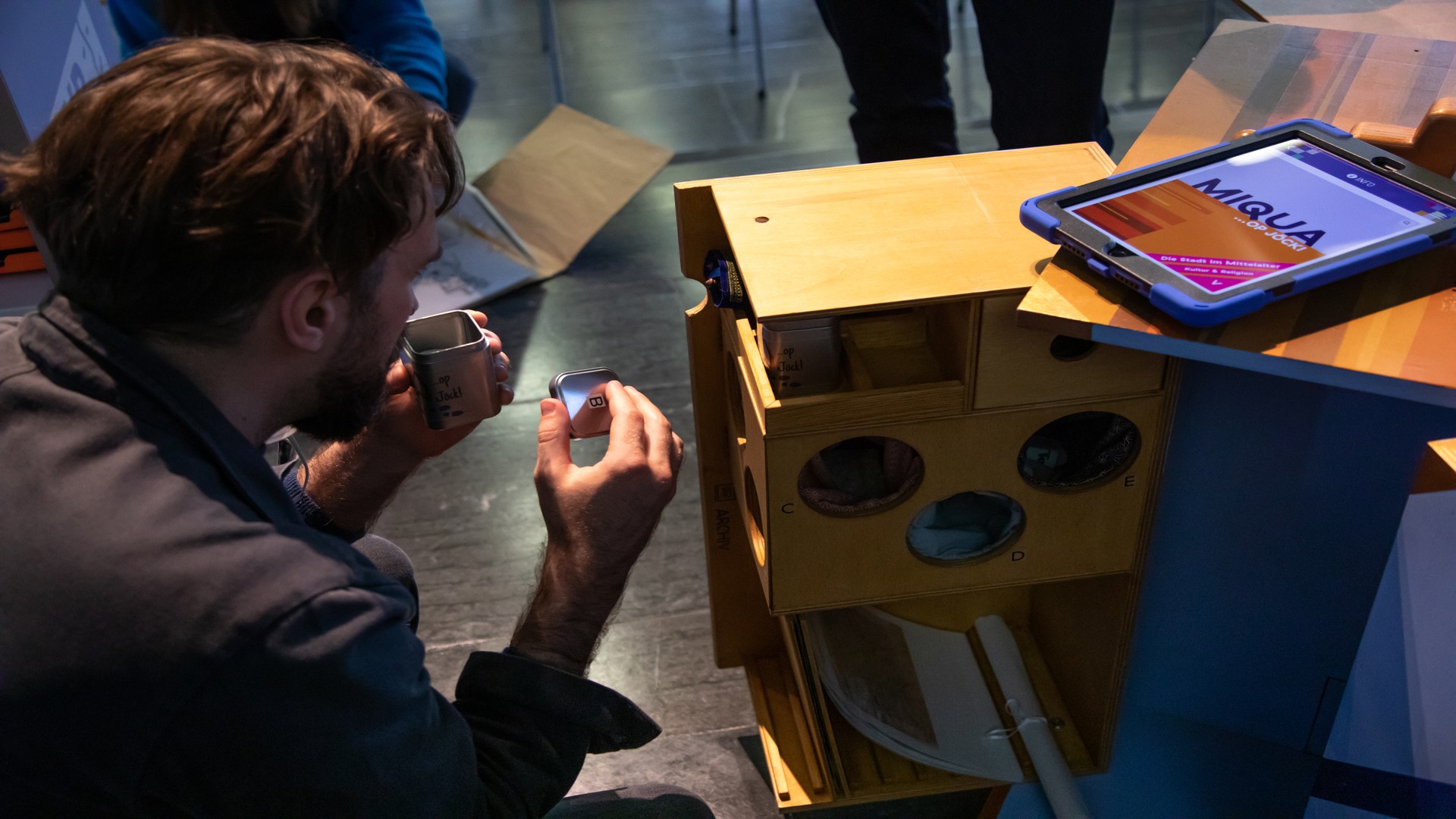 Ein Mann hockt vor dem mobilen Möbel "MiQua...op Jöck!", das ausgefaltet in einem Raum steht. Auf dem mobilen Möbel liegt ein Ipad, auf dem die "MiQua...op Jöck!" angezeigt wird. Der Mann riecht an einer kleinen Dose.