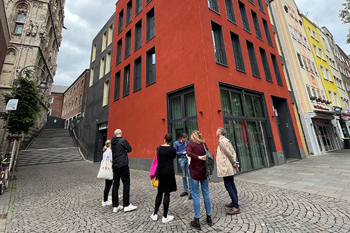 Eine Gruppe Erwachsener steht vor dem roten Haus am Alter Markt, wo das MiQua:forum situiert ist.  Sie hören einem Stadtführer zu.