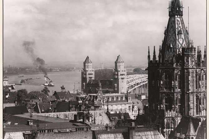 Schwarz-weiß Foto des Kölner Rathausturms mit Blick auf den Rhein, um 1938. Auf dem Rhein fährt ein dampfendes Schiff.